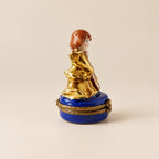 A small figurine of a seated woman with reddish-brown hair, gold clothing, and a blue base with gold trim, displayed on a white circular platform against a light gray background.
