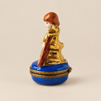 A decorative domed jewelry box featuring a porcelain figurine of a woman with reddish-brown hair and a gold dress, seated on a glossy blue base with ornate gold trim.