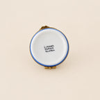 A vintage French enamel pin by Ladybird Laugh, featuring a white enamel face with a blue rim, photographed from the back on a white circular display stand against a neutral gray background.