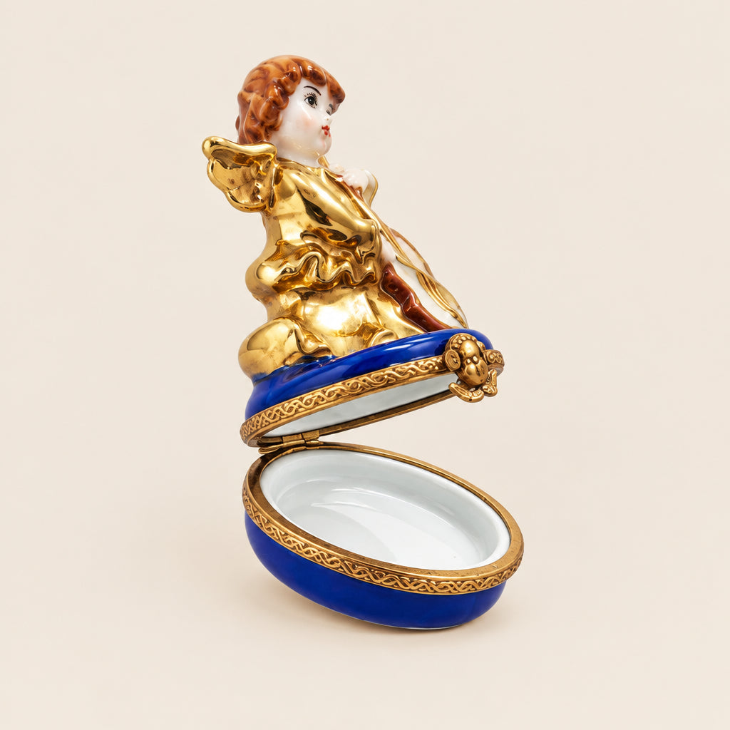 A small decorative figurine of a cherub or angel posed with one leg raised, mounted on a round white display base against a plain light gray background. The figure is primarily gold with blue trim and a reddish-brown hair accent.