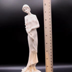White ceramic figurine of a woman with a flower, next to a ruler for scale, on a dark background.