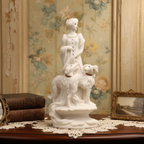 White marble sculpture of a woman with two dogs on a wooden table with floral wallpaper.