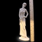 White ceramic figurine of a woman with a hat next to a ruler for scale on a black background