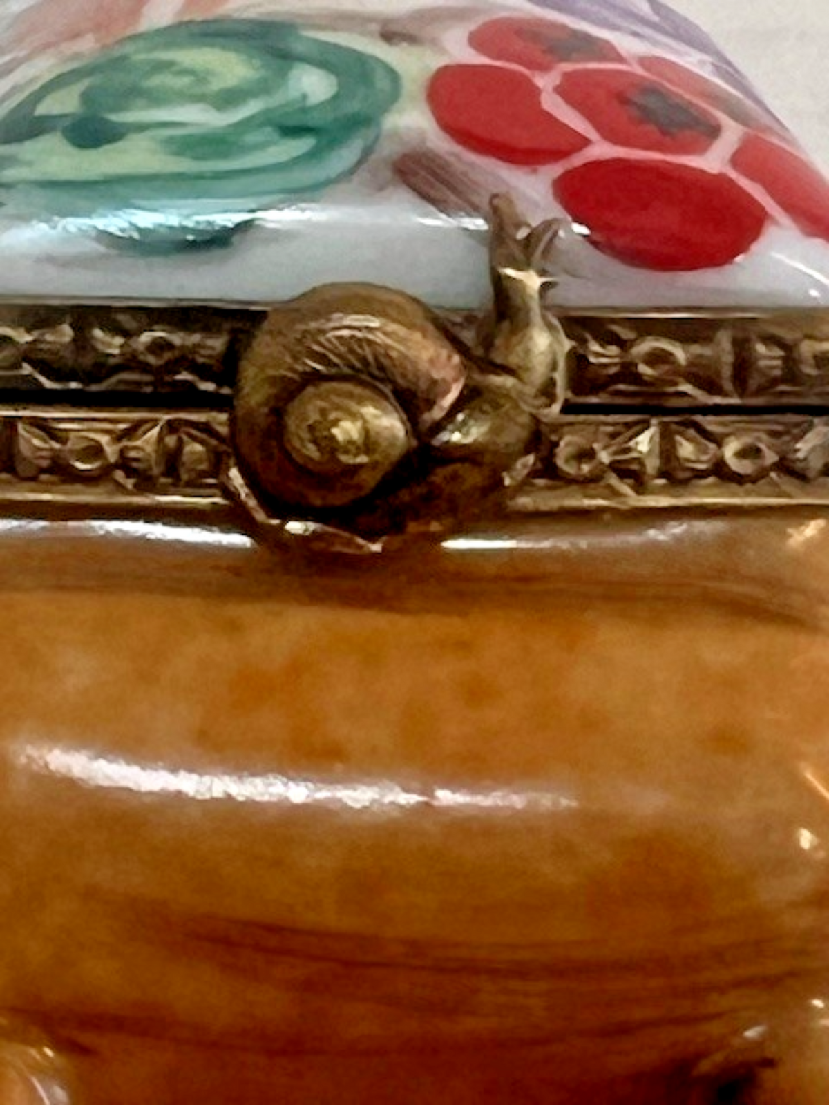Close-up of a decorative ceramic or porcelain container lid featuring a gold-toned snail clasp, floral painted details in red and green, and a glossy warm brown body.